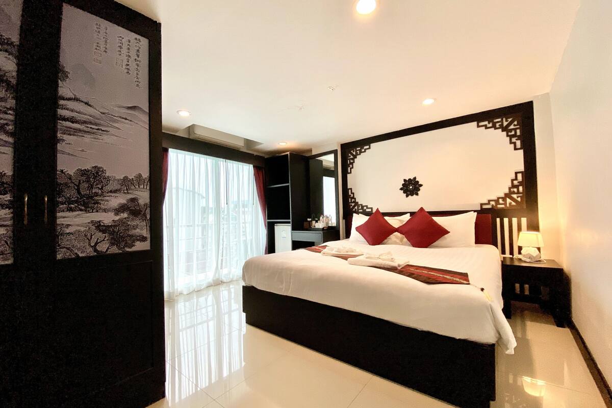 A modern hotel room with a large bed, a nightstand, a wall-mounted TV, and a large window with sheer curtains.