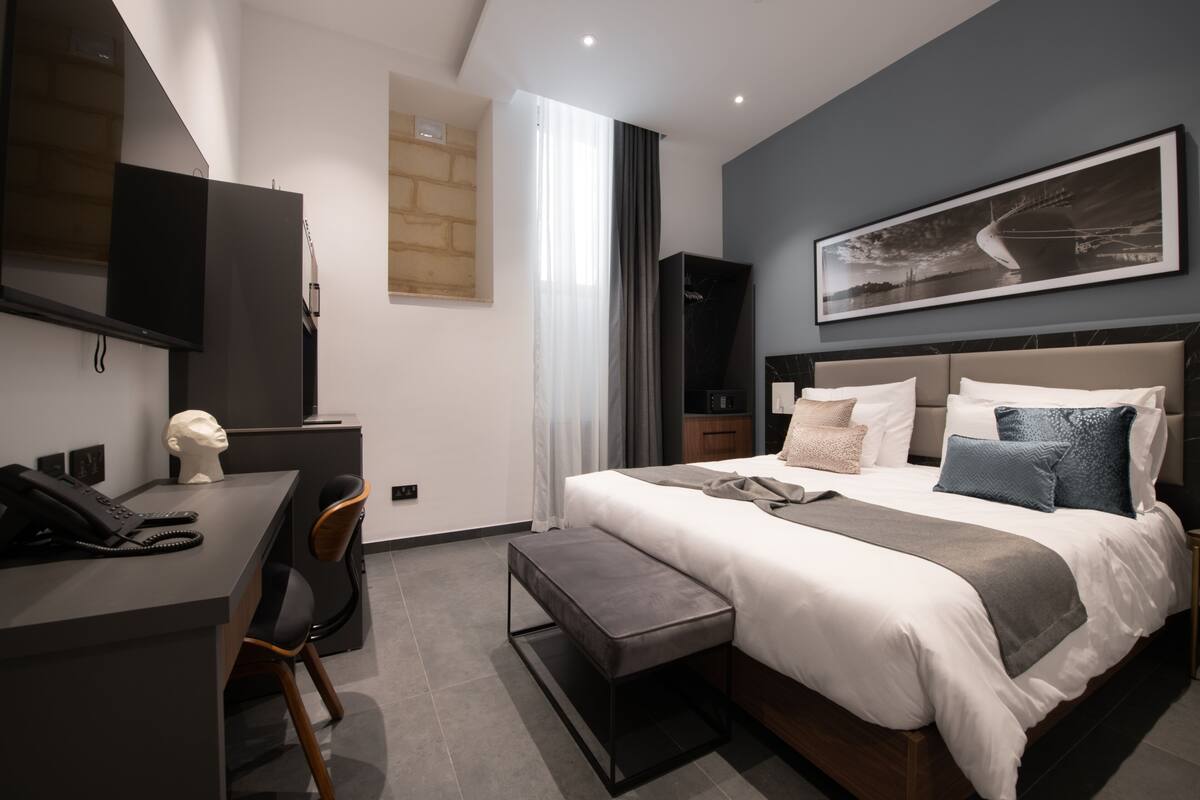 A modern hotel room with a large bed, a desk with a telephone, a chair, and a bench.