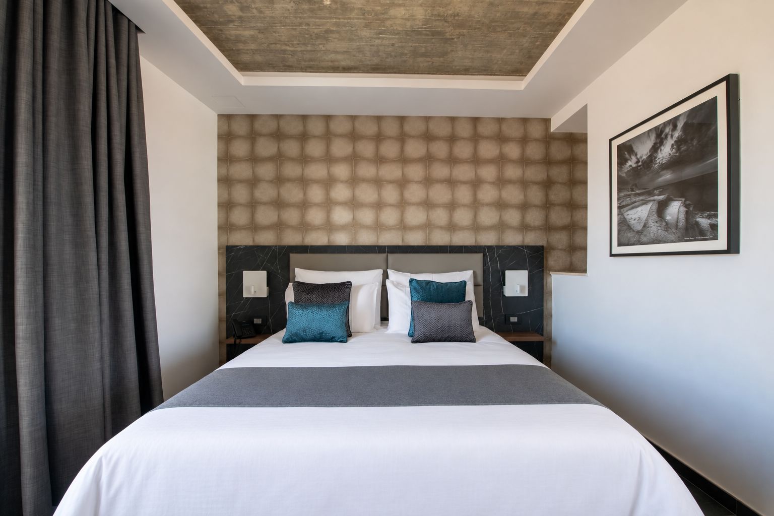 Une chambre d’hôtel avec un grand lit, une tête de lit à motifs, un tableau encadré au mur et un rideau foncé.