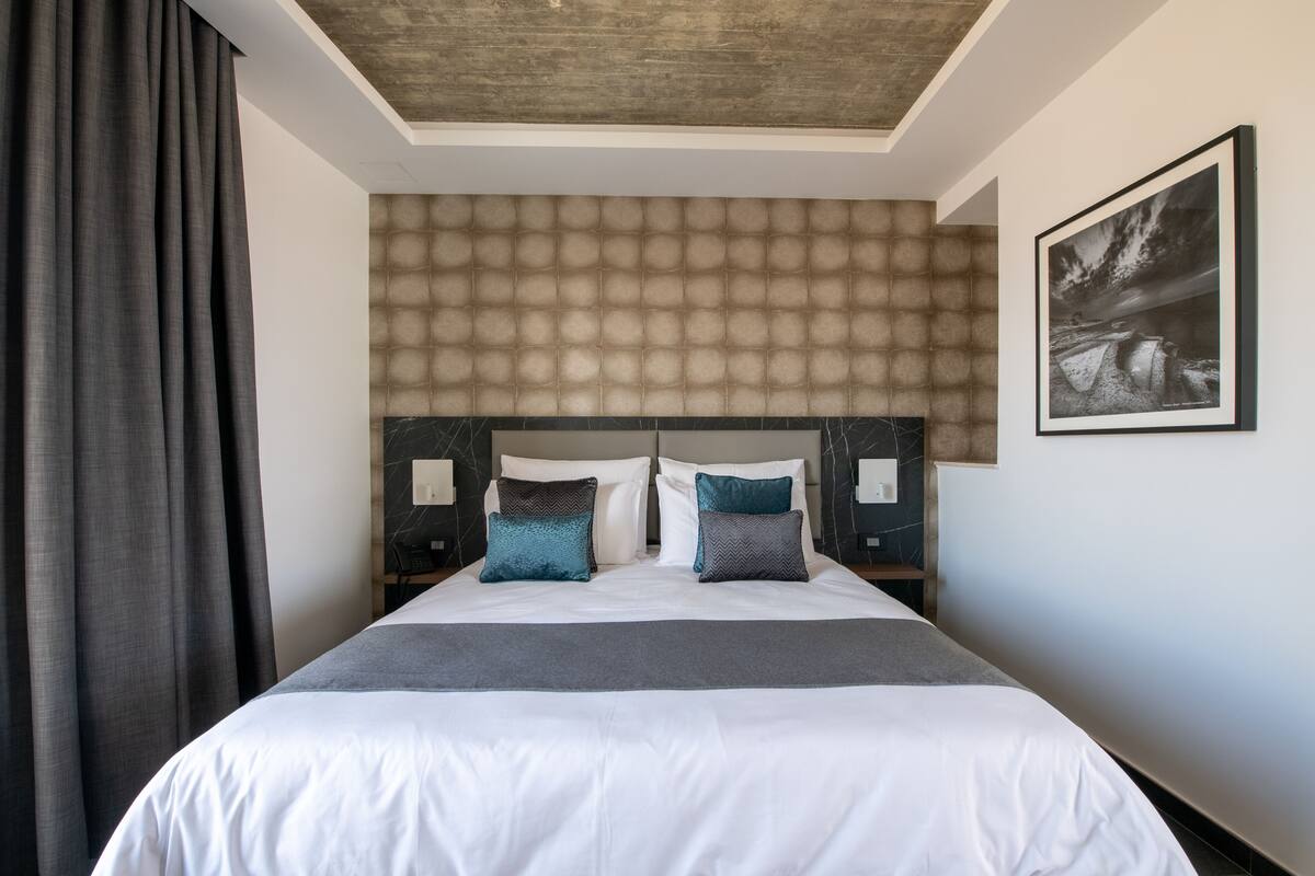 A modern hotel room with a large bed, a headboard with a textured pattern, two bedside tables with lamps, and a framed picture on the wall.