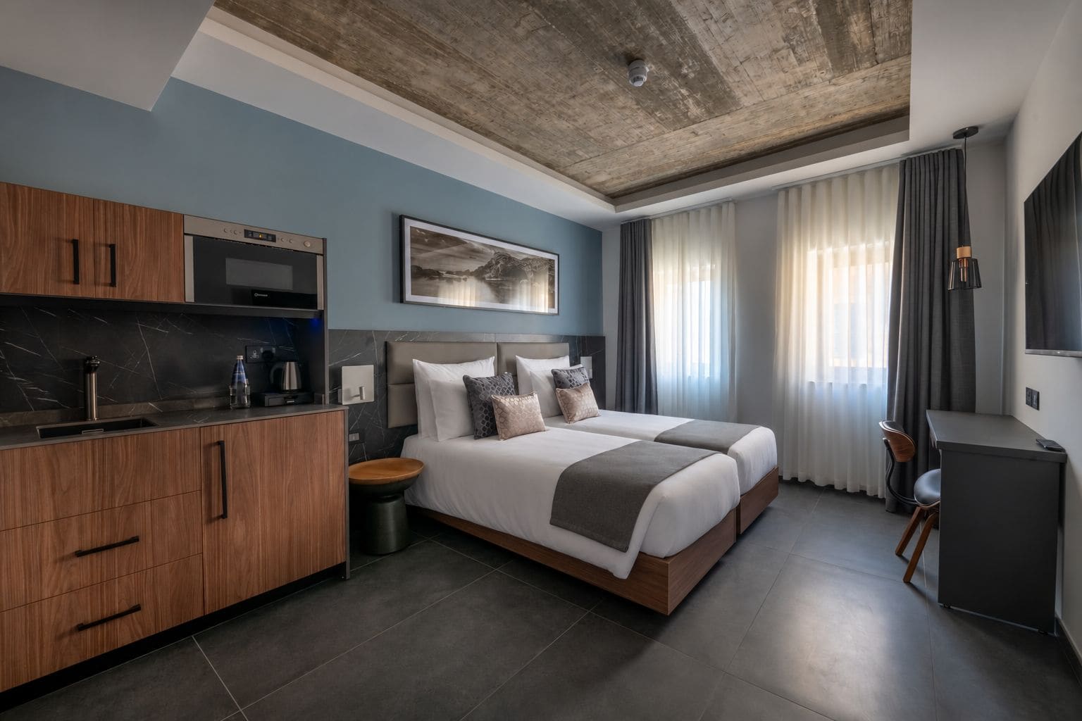 A modern hotel room with a kitchenette, a large bed with multiple pillows, a desk, and a large window with curtains.