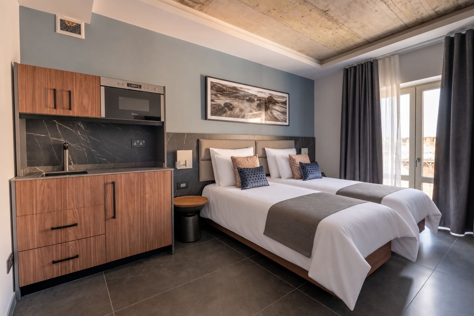 A hotel room with a kitchenette, two beds, a large window with curtains, and a painting on the wall.