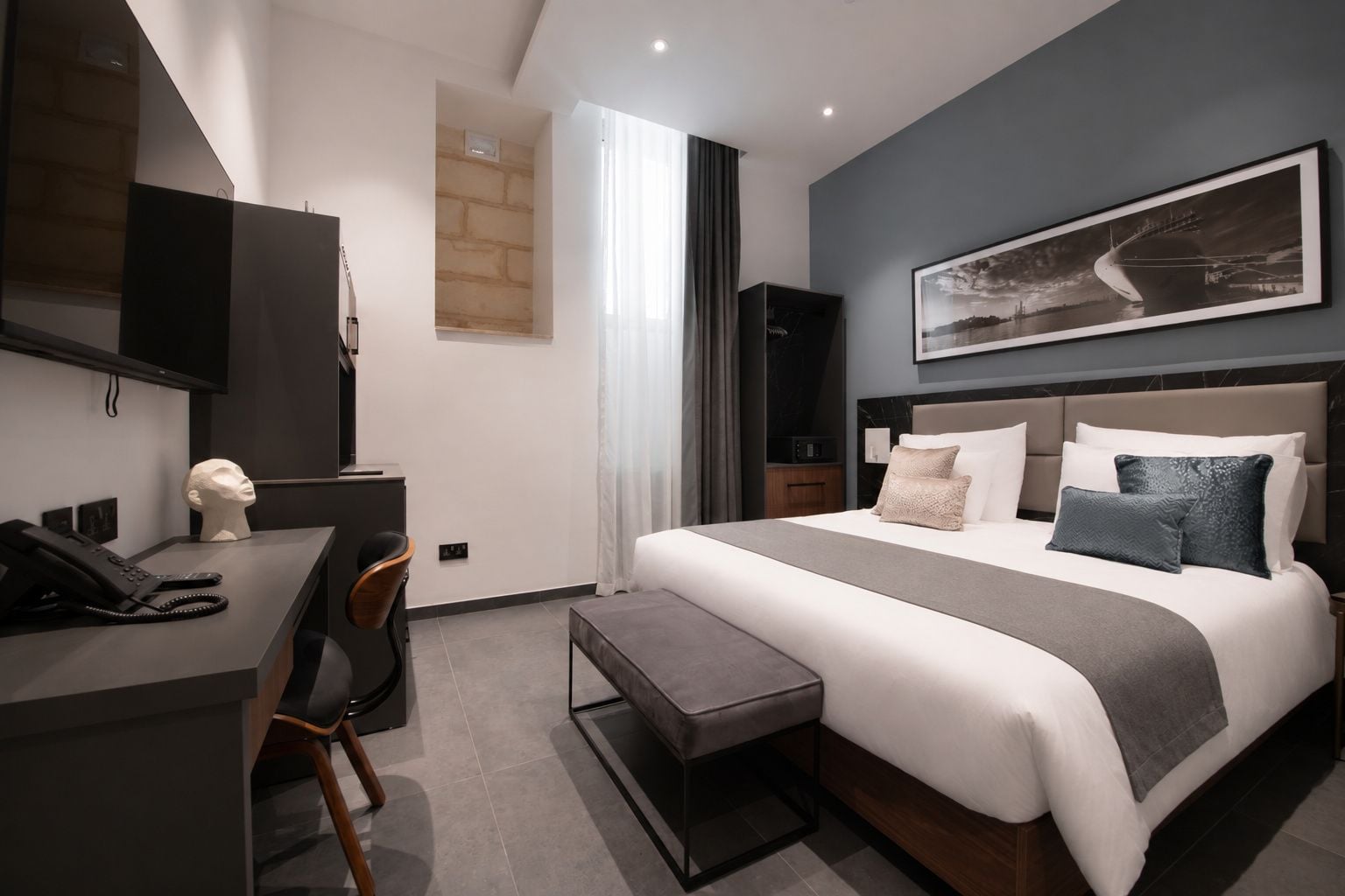 A modern hotel room with a bed, desk, chair, and a mounted TV.