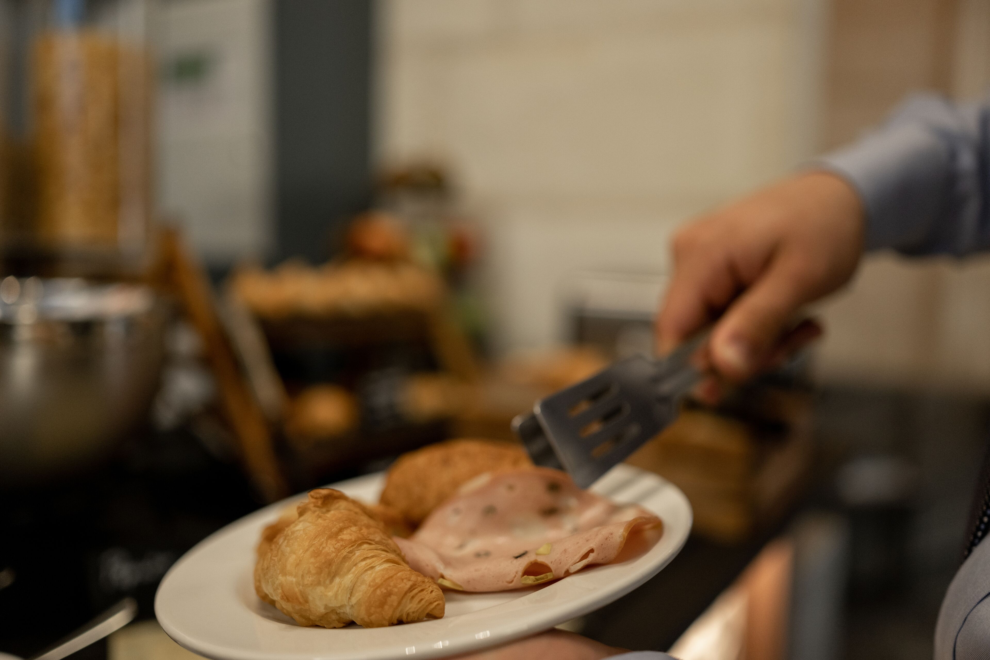 Ein Teller mit Croissants und Schinken, eine Person hält einen Pfannenwender, und ein unscharfer Hintergrund mit Küchenutensilien.