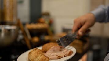 Ein Teller mit Croissants und Schinken, eine Person hält einen Pfannenwender, und ein unscharfer Hintergrund mit Küchenutensilien.