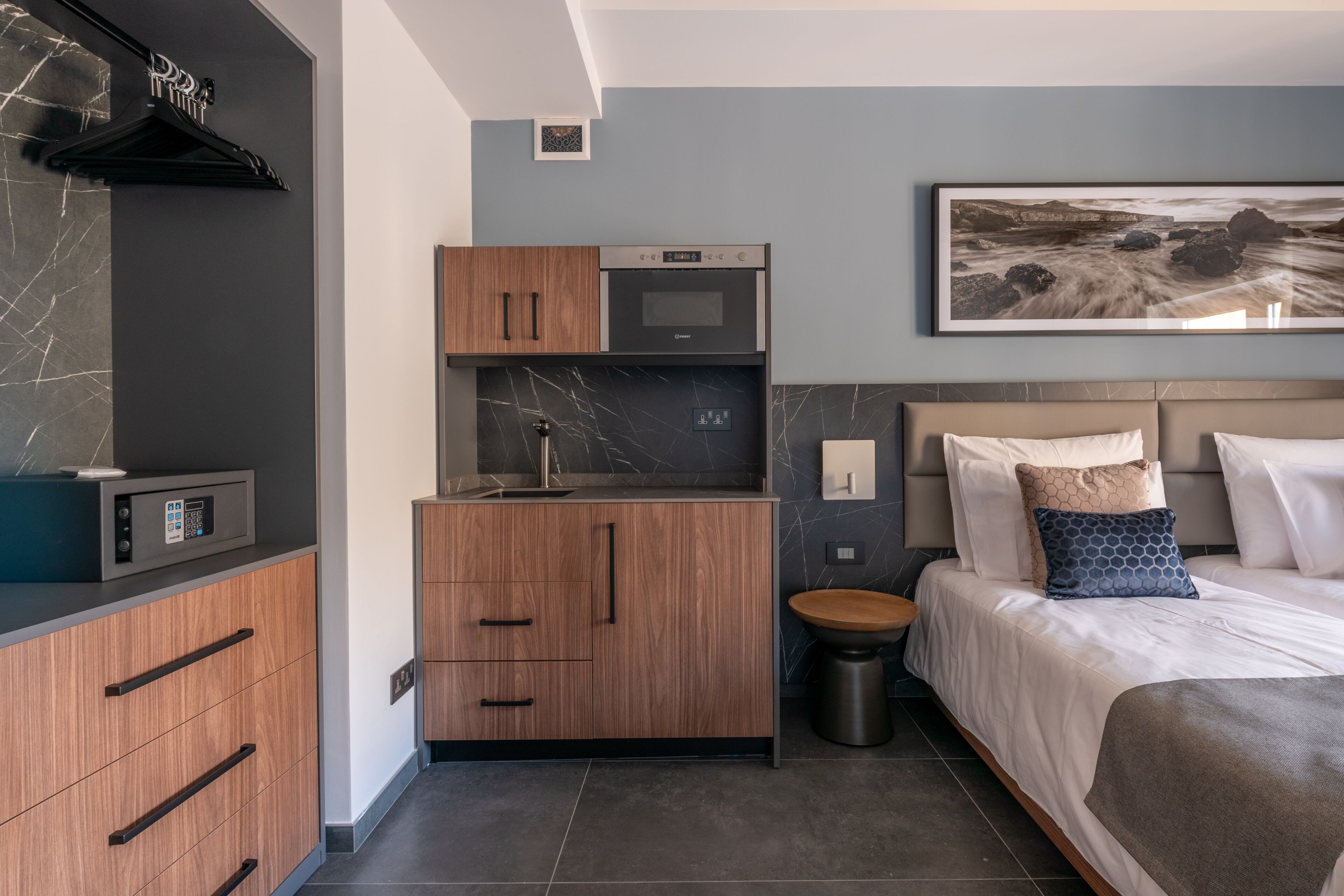 A compact hotel room with a kitchenette, a bed with a headboard, and a painting on the wall.