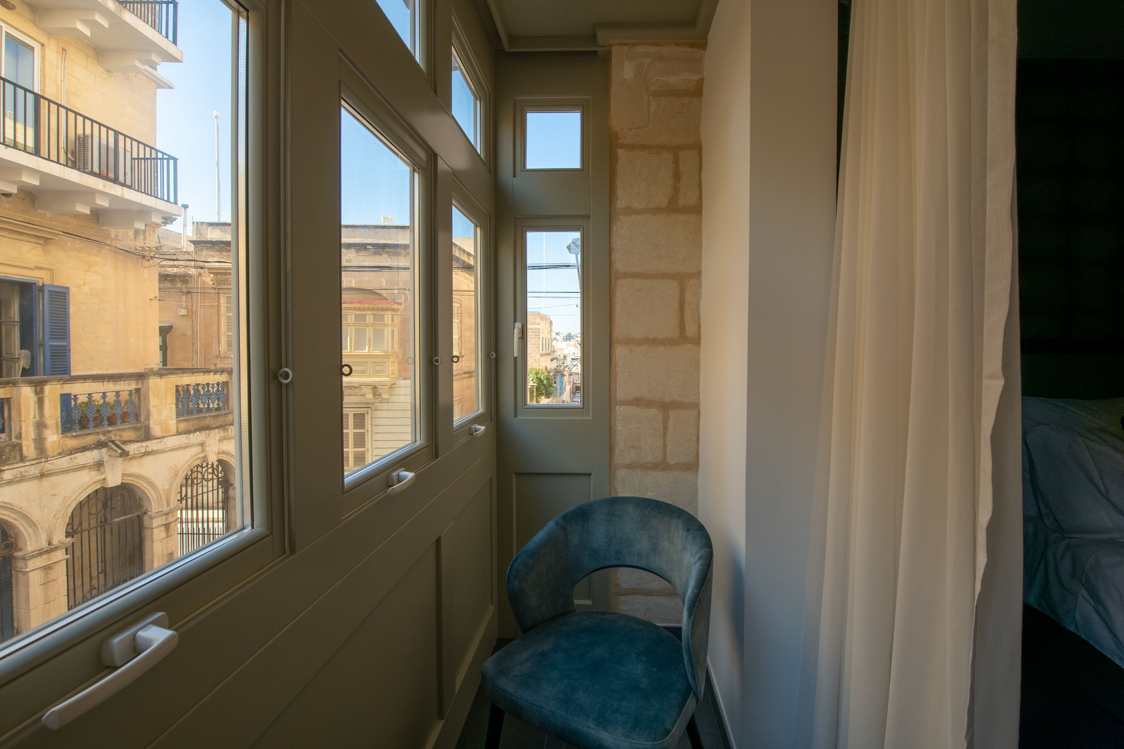 A narrow hallway with a chair, a bed, and a view of a building with balconies.