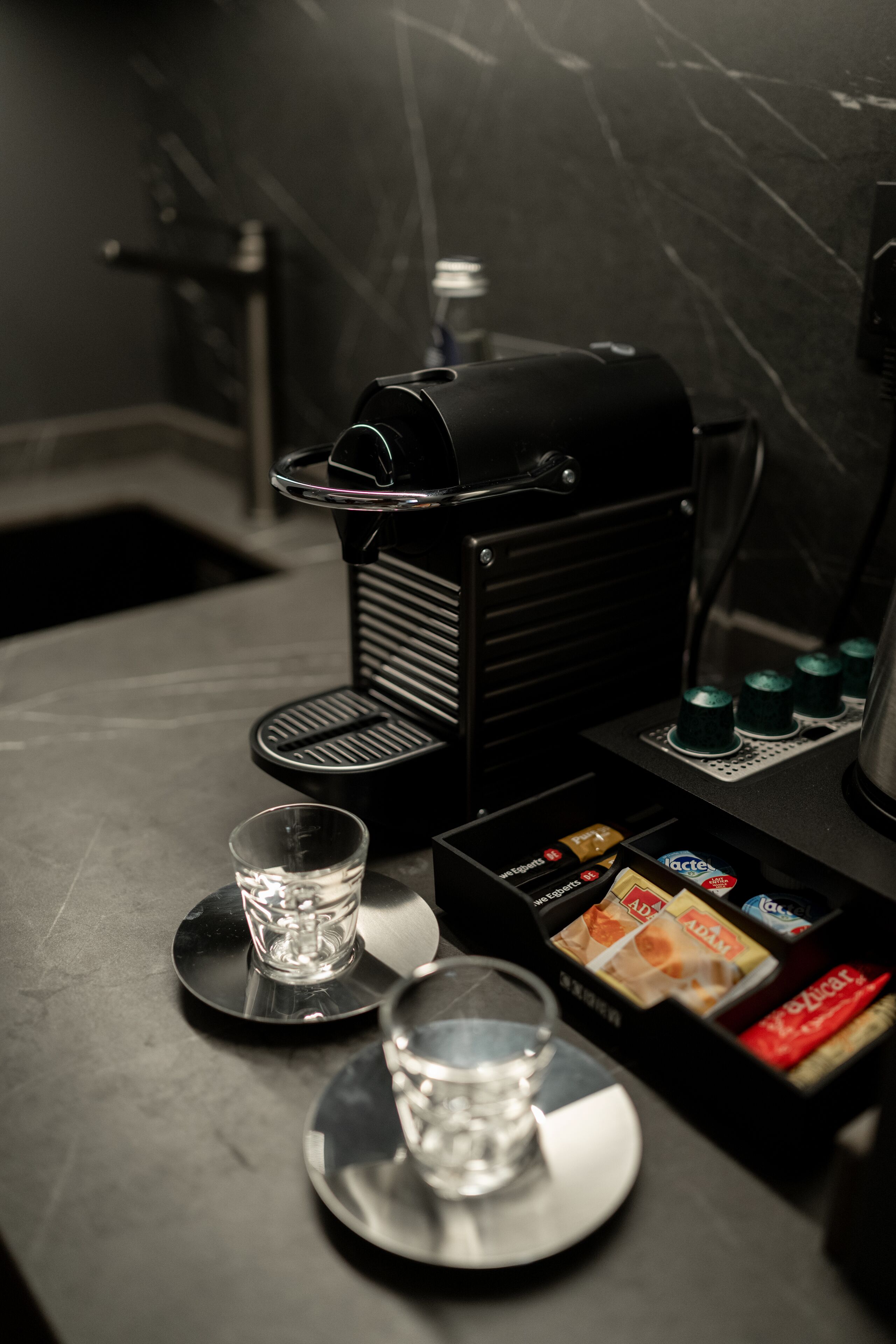 Een koffieapparaat met een waterreservoir, een glas en koffiekapsules op een aanrecht.