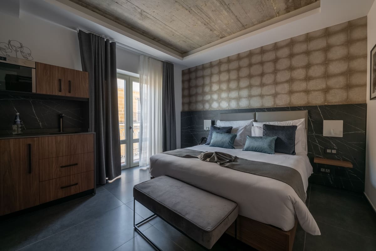 A modern hotel room with a large bed, a wooden wardrobe, a bench, and a view of the outdoors.