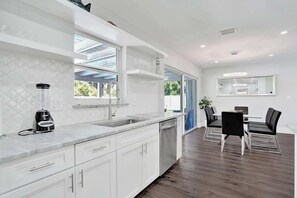 A modern kitchen with white cabinetry, a built-in dishwasher, and a dining area with a table and chairs. - Gorgeous 4BR| Brand new HTD Pool & Jacuzzi| 1Mile (Deerfield Beach)