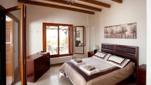 Een slaapkamer met een groot bed, houten balken in het plafond, een schuifdeur naar een terras, en een lijst met bloemistisch kunstwerk aan de muur.