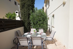 Eine Terrasse mit einem für vier Personen gedeckten Tisch, umgeben von Stühlen, mit einem hohen Baum und einem Gebäude im Hintergrund.