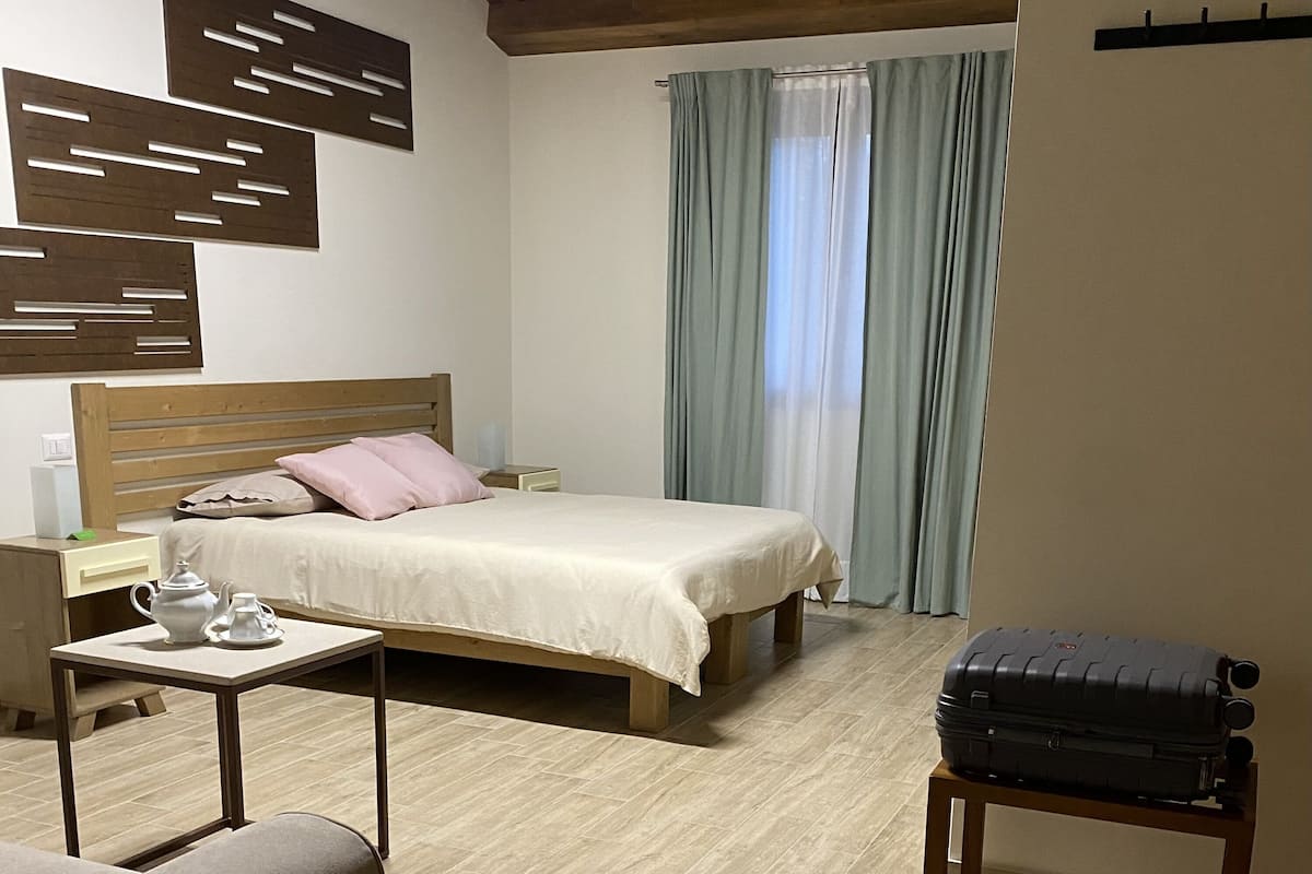 Un dormitorio con cama, mesita de noche, lámpara, cortinas y una maleta.