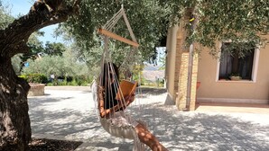 Une personne se détend dans un hamac sous un arbre, avec une terrasse pavée et un bâtiment en arrière-plan.