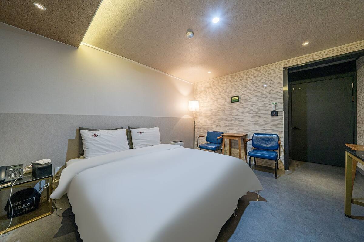 Una habitación de hotel con una cama grande, mesita de noche, lámpara y dos sillones azules.