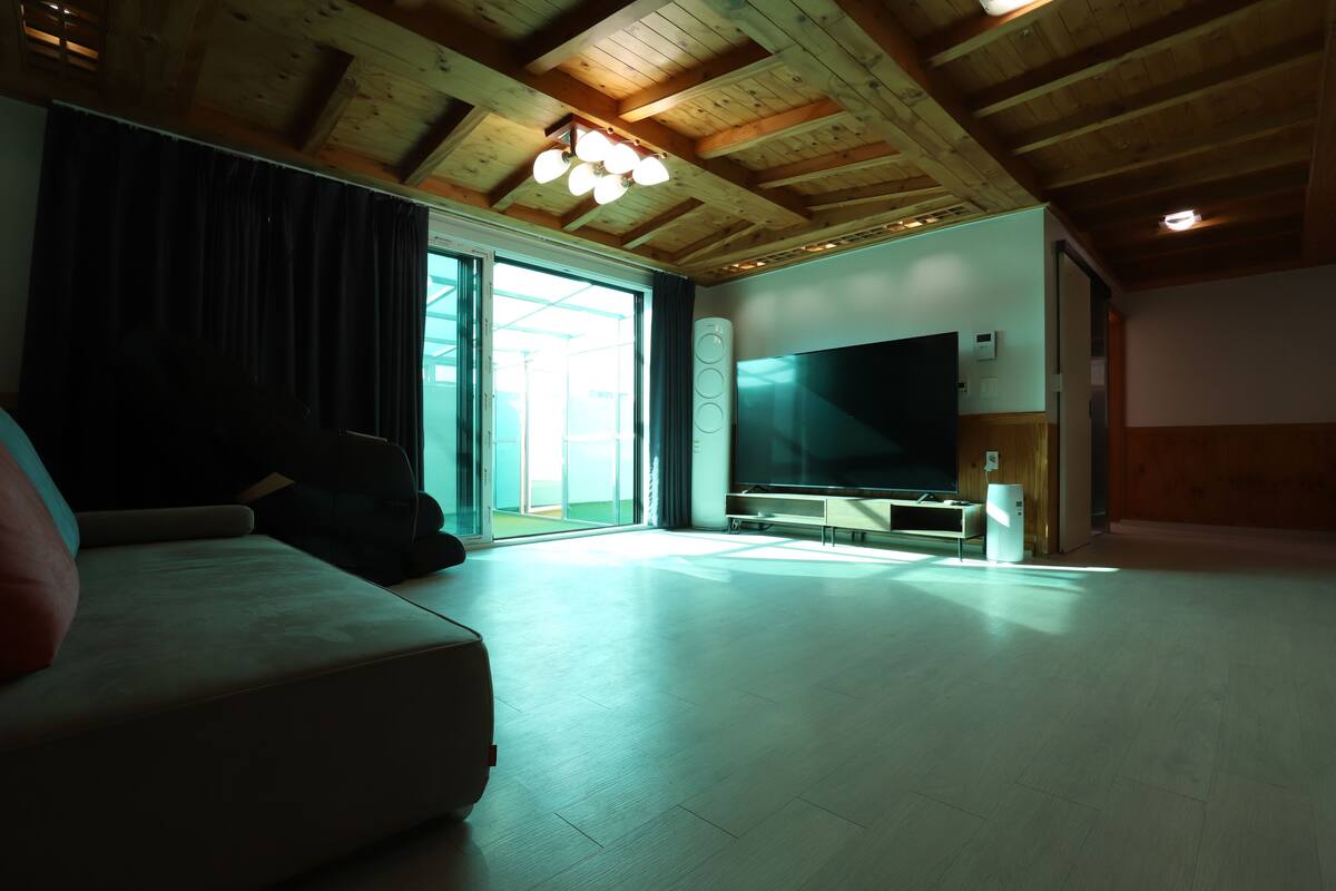 Un salón espacioso con una televisión de pantalla plana grande, vigas de madera en el techo y un gran ventanal corredero que da a un espacio exterior.