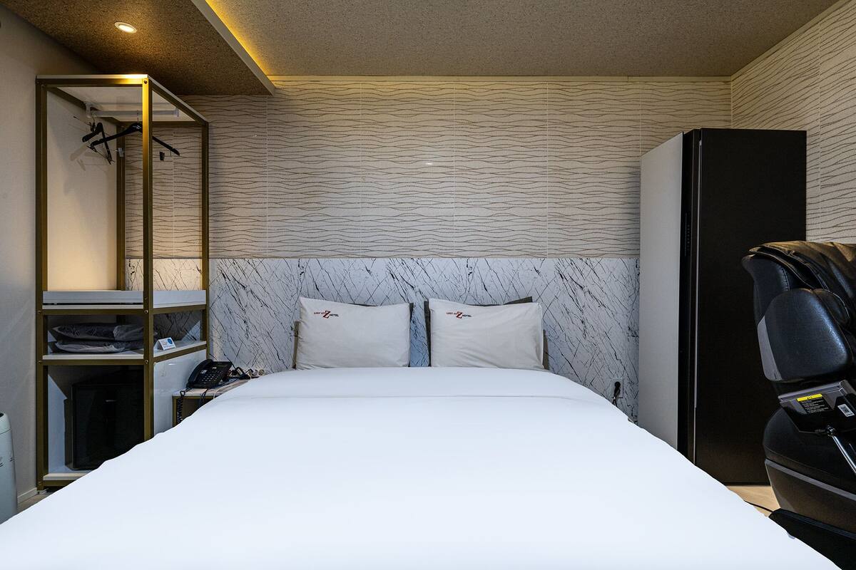 Habitación de hotel con cama, armario, estante con ropa y sillón de masaje.