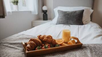 En snyggt bäddad säng med ett brickbord som innehåller croissanter, jordgubbar och apelsinskivor, och ett glas apelsinjuice.