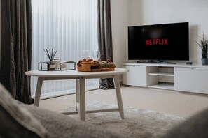 Ein modernes Wohnzimmer mit einem Flachbildfernseher, der das Netflix-Logo anzeigt, einem runden Tisch mit Obstschale und Kerzen sowie einem großen Fenster mit transparenten Vorhängen.