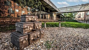 Een stapel oude koffers op een grindpad, met een bakstenen gebouw en een voetgangersbrug op de achtergrond.