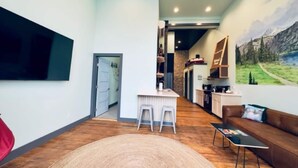 Una sala de estar moderna con un televisor grande, un tapete redondo, un sofá y una cocina visible a través de una puerta abierta.
