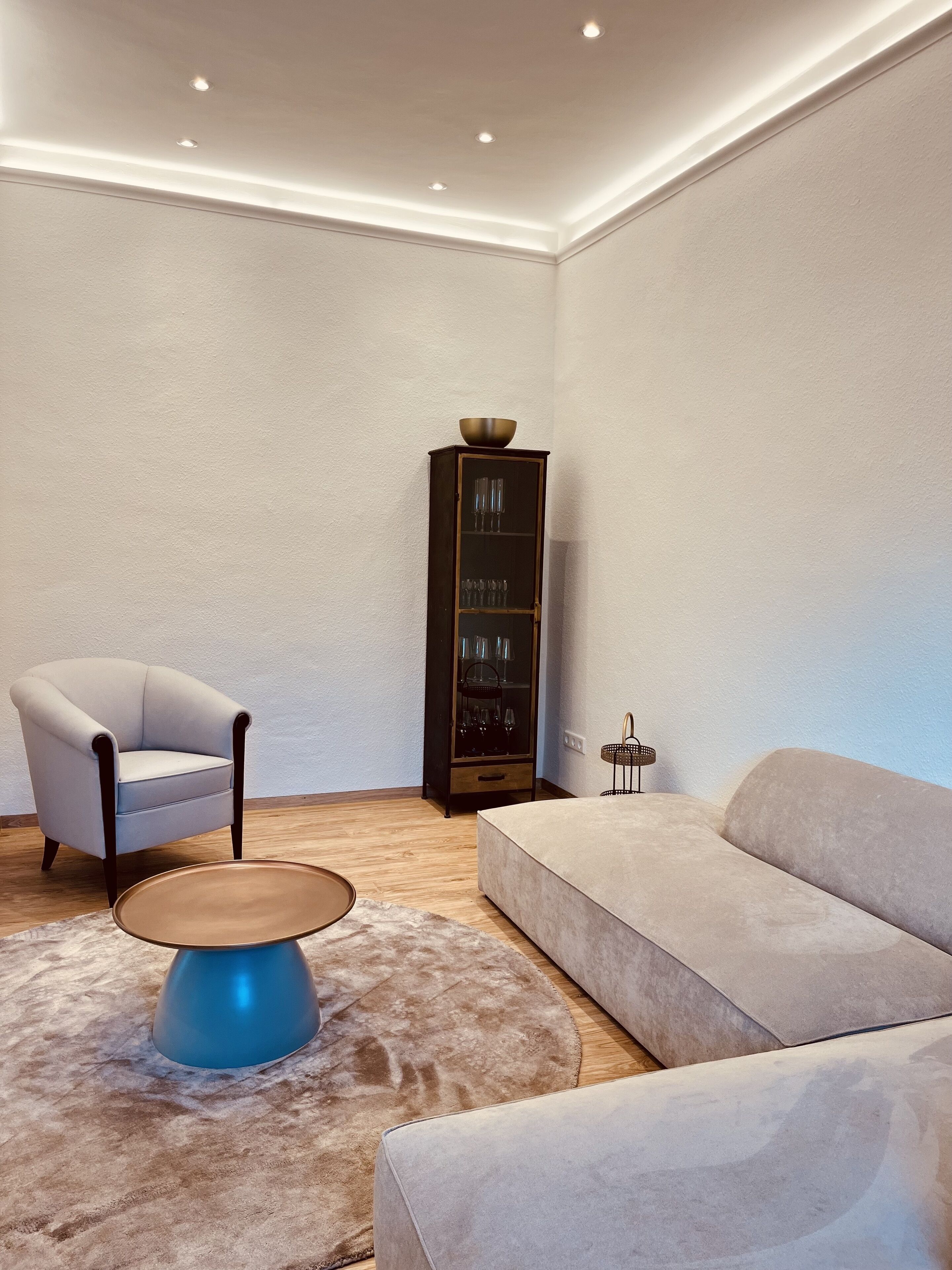 A modern living room with a light grey sofa, a round coffee table, a grey armchair, and a dark wooden cabinet with glass doors.