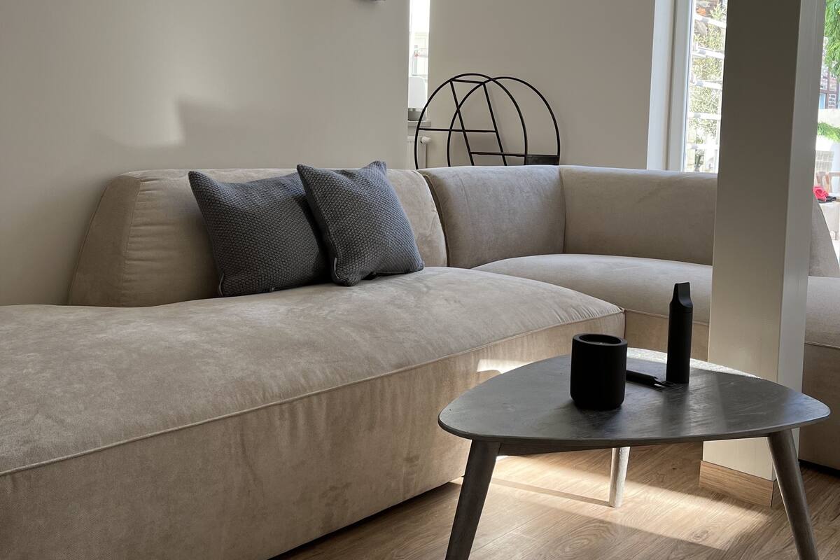 Una sala moderna con un sofá seccional color beige, dos almohadas grises, una mesita redonda con un jarrón negro y una vela, y piso de madera.
