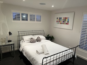 Cama bem arrumada com edredom branco e almofadas, mesa de cabeceira com abajur, e paleta de cores na parede.
