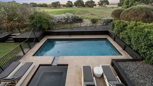 A rectangular swimming pool with a tiled deck, surrounded by a black metal fence. There are lounge chairs and a small round table near the pool. - Beach house on a golf course (Torquay)