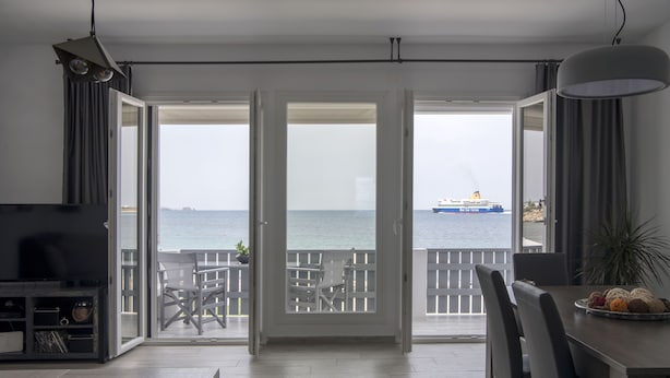 A room with a large sliding glass door leading to a balcony with a view of the sea and a ship. - Parikia's Seaside House (Paros)