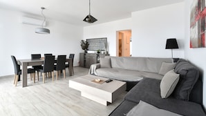 A modern living room with a grey sofa, a dining table with chairs, a coffee table, and a kitchen in the background. - Parikia's Seaside House (Paros)