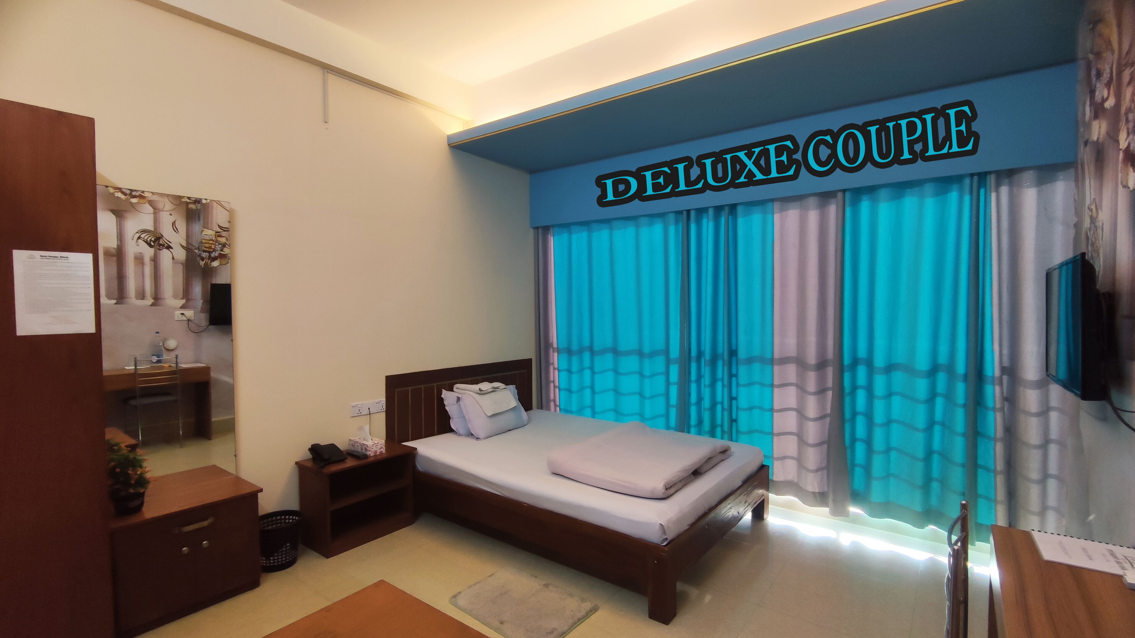 A hotel room with a bed, a desk, a chair, a TV, and a sign that reads "DELUXE COUPLE".