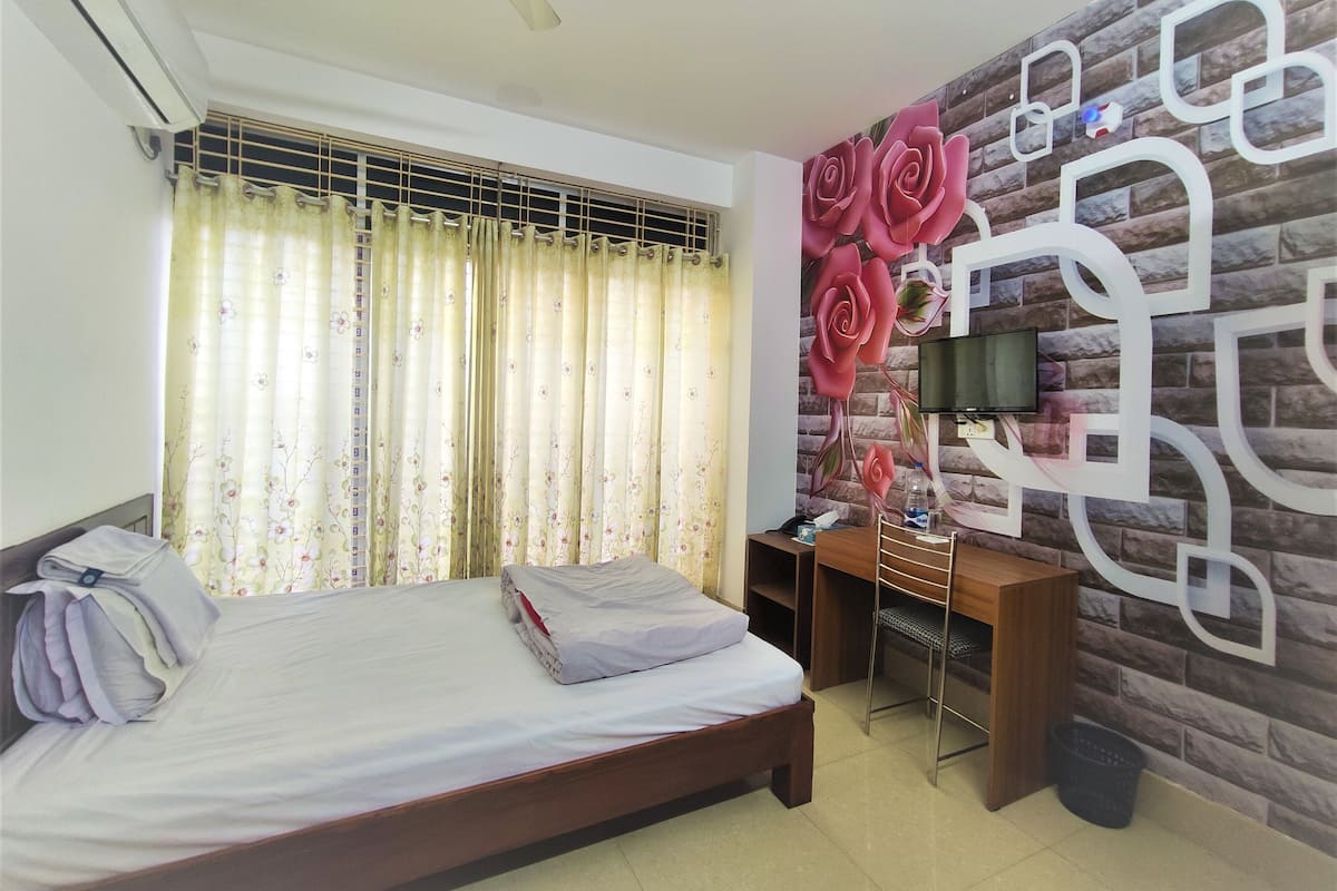 A hotel room with a bed, a desk with a chair, a TV mounted on a brick-patterned wall, and a window with floral curtains.