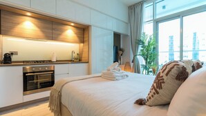 A modern hotel room with a kitchenette, a large bed with white linens, and a view of the city through the window. - Stunning Apt Marina view with Balcony (Dubai)