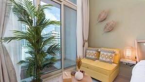 A modern hotel room with a large window, a yellow sofa, a small round table, and a potted plant. - Stunning Apt Marina view with Balcony (Dubai)