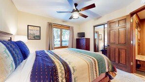 Un dormitorio con una cama, un ventilador de techo, un televisor sobre un mueble de madera y una puerta que da a otra habitación.