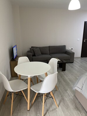 Moderni olohuone, jossa on harmaa sohva, pyöreä valkoinen pöytä ja neljä tuolia sekä pieni puinen sivupöytä, jonka päällä on televisio.