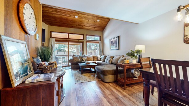 A cozy living room with a wooden ceiling, large windows, a sofa, a coffee table, and a dining table. - Woodlands #11 3 Bedroom Condo by RedAwning (Mammoth Lakes)