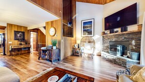 A spacious living room with a wooden floor, a fireplace, a large TV, and a dining area with a wooden table and chairs. - Woodlands #11 3 Bedroom Condo by RedAwning (Mammoth Lakes)