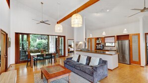 Uma sala de estar ampla com sofá, mesa de centro de madeira e iluminação pendente. Cozinha com geladeira e ilha é visível ao fundo.