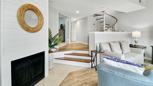 A modern living room with a fireplace, a round woven mirror, a staircase with a wooden railing, and a sofa with cushions. - Updated 3 bedroom Villa in Shelter Cove with Community Pool (Hilton Head Island)