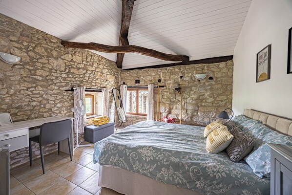 A bedroom with a stone wall, a bed with patterned bedding, a desk with a chair, and a window with curtains.