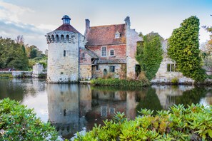A historic stone building with a tower, a red-roofed main structure, and a reflection in the water.