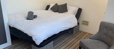 A neatly made bed with white bedding and gray pillows, a gray upholstered armchair, and a striped carpet.