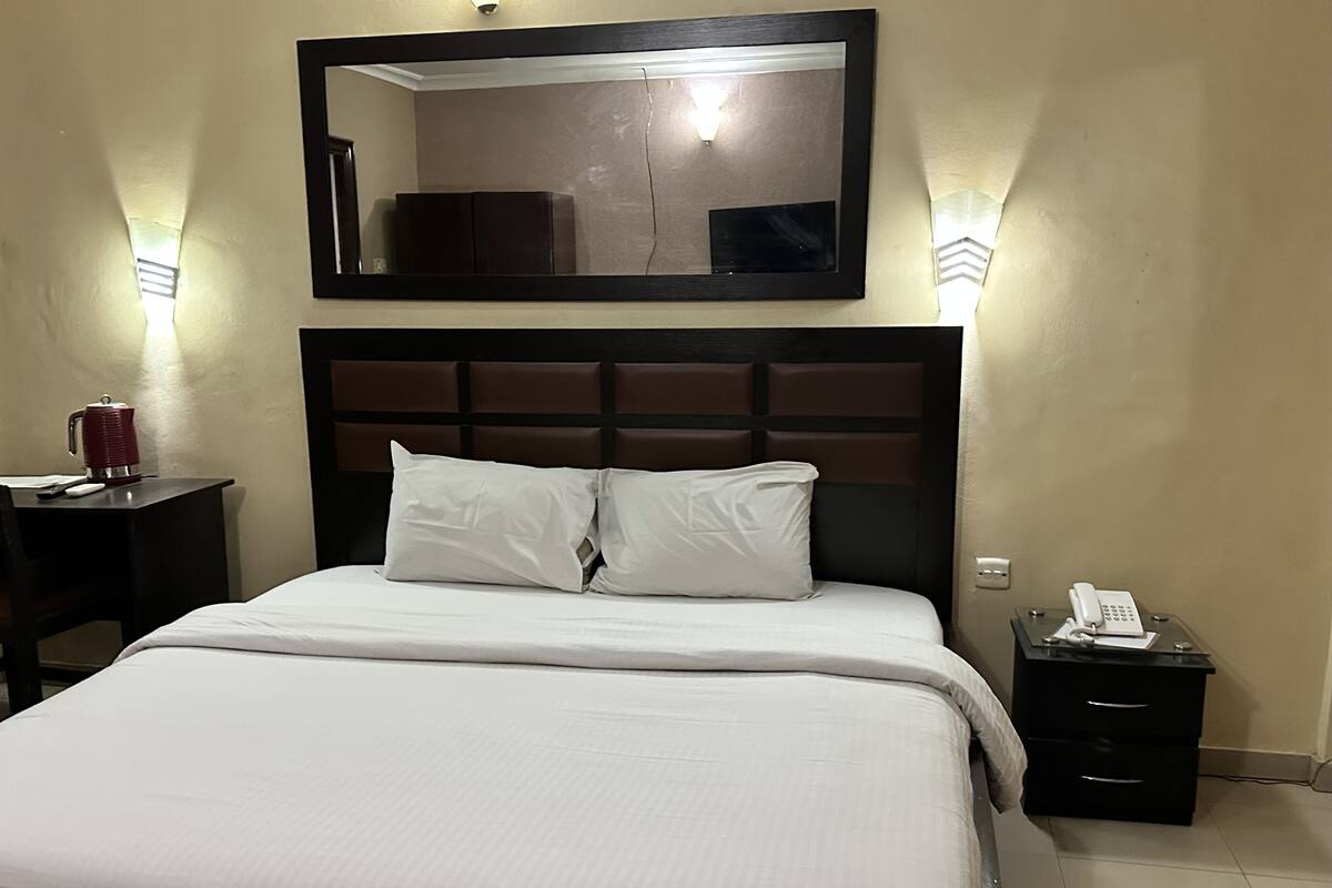 A hotel room with a bed, a nightstand with a phone, a mirror above the bed, and wall sconces.