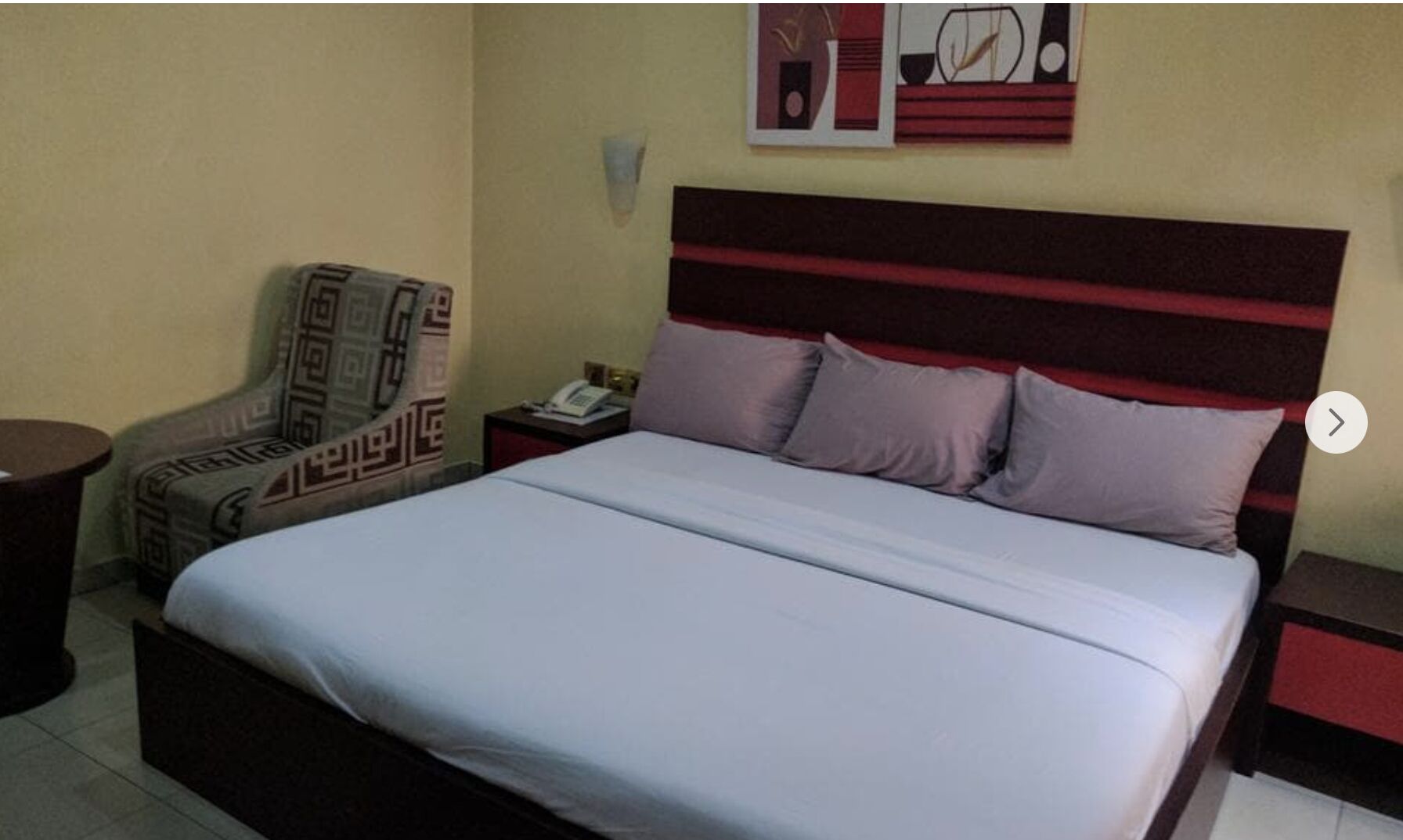 A hotel room with a bed, a chair, a nightstand, and a wall with artwork.