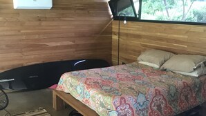 Una camera da letto con soffitto in legno, un letto con biancheria a motivi, una panca, una sedia e una finestra con vista sugli alberi.