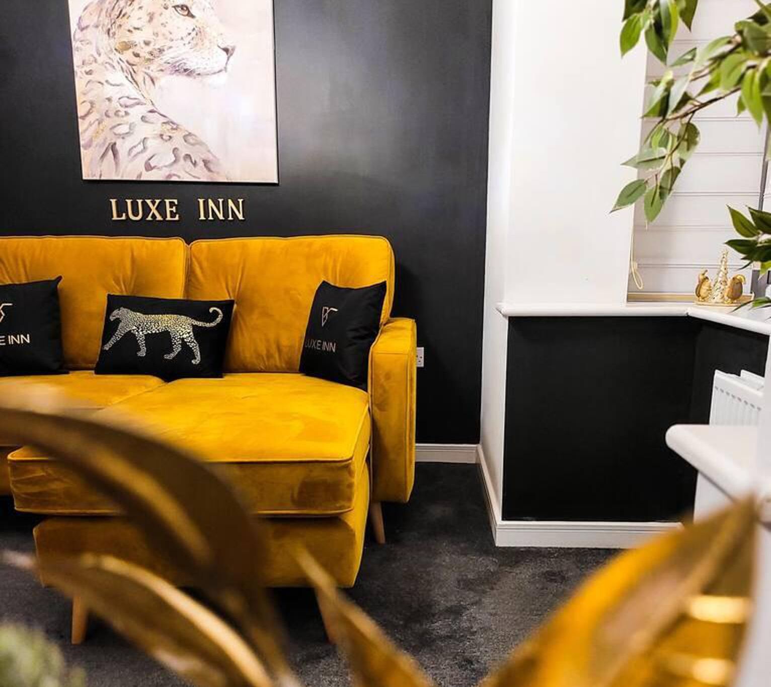 Ein Zimmer mit einem gelben Sofa, schwarzen Kissen, einem leopardenmuster-Kissen und einer schwarz-weißen Wand mit einem 'LUXE INN' Schild.