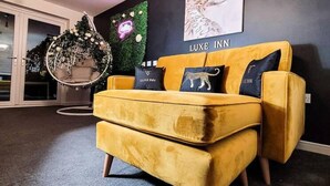 Ein gelbes Sofa mit 'LUXE INN'-Branding, eine hängende Leuchte, ein Wandbild von einem Hund und eine grüne Wanddekoration.