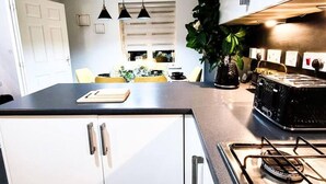Eine moderne Küche mit weißen Schränken, einer schwarzen Arbeitsplatte und einem Gasherd. Über der Kochinsel hängen Pendelleuchten und auf der Arbeitsfläche steht ein Blumentopf.